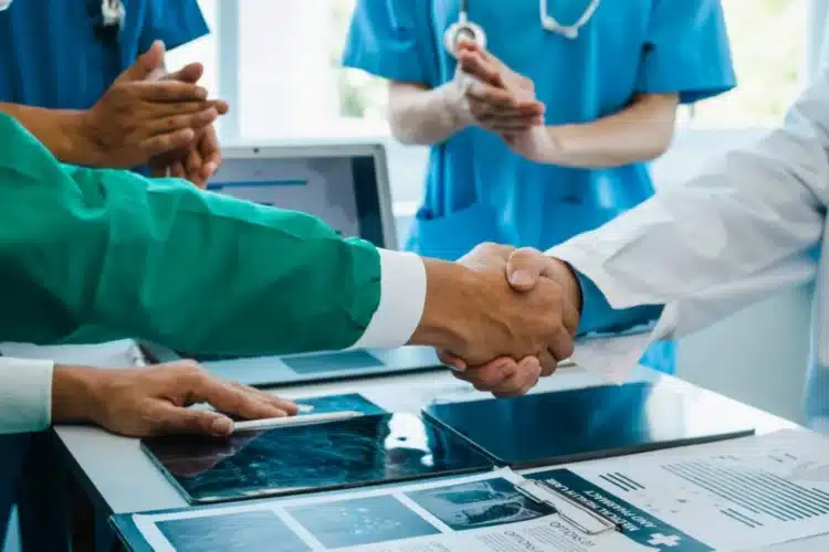 Doctors in a healthcare setting shaking hands and collaborating on patient care with medical reports and technology on the table.