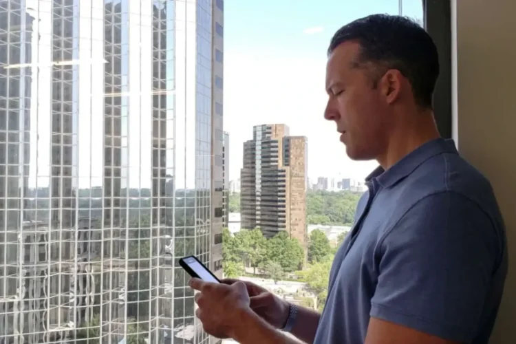 A businessman standing by a window overlooking city buildings, using a smartphone to explore property listings, symbolizing real estate app development for modern buyers and sellers.