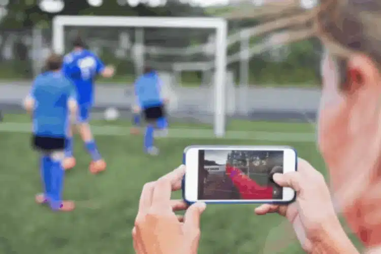 A person is holding a smartphone, recording a soccer match, with a digital overlay showing player statistics or analysis on the screen, illustrating Javascript sports app development.