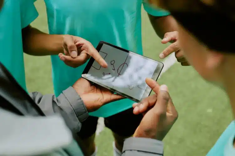 A group of athletes and coaches huddle around a tablet, pointing at a sports play diagram, an example of sports data visualization UI in action.