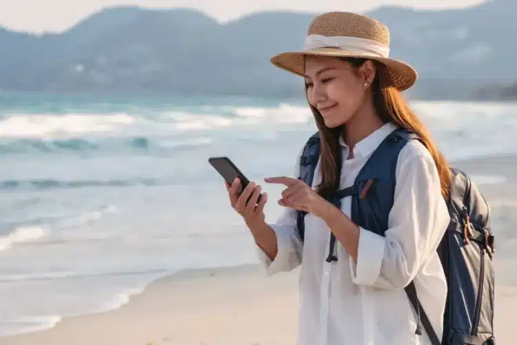 A traveler with a backpack using a smartphone on the beach, symbolizing travel app development that enhances trip planning, bookings, and navigation for tourists.