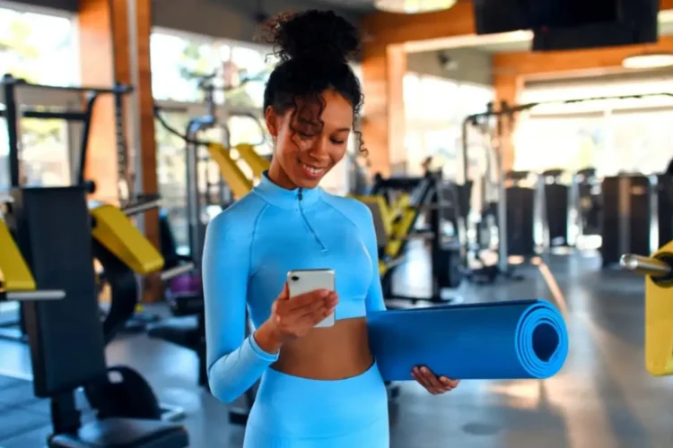 A woman in activewear holding a yoga mat and checking her smartphone in a gym, representing fitness app development for workout tracking and personalized training plans.