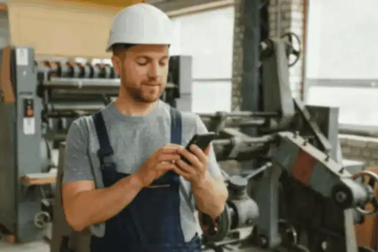 A male manufacturing worker wearing a hard hat and overalls, using a smartphone in a factory, representing the application of React Native in manufacturing app development.