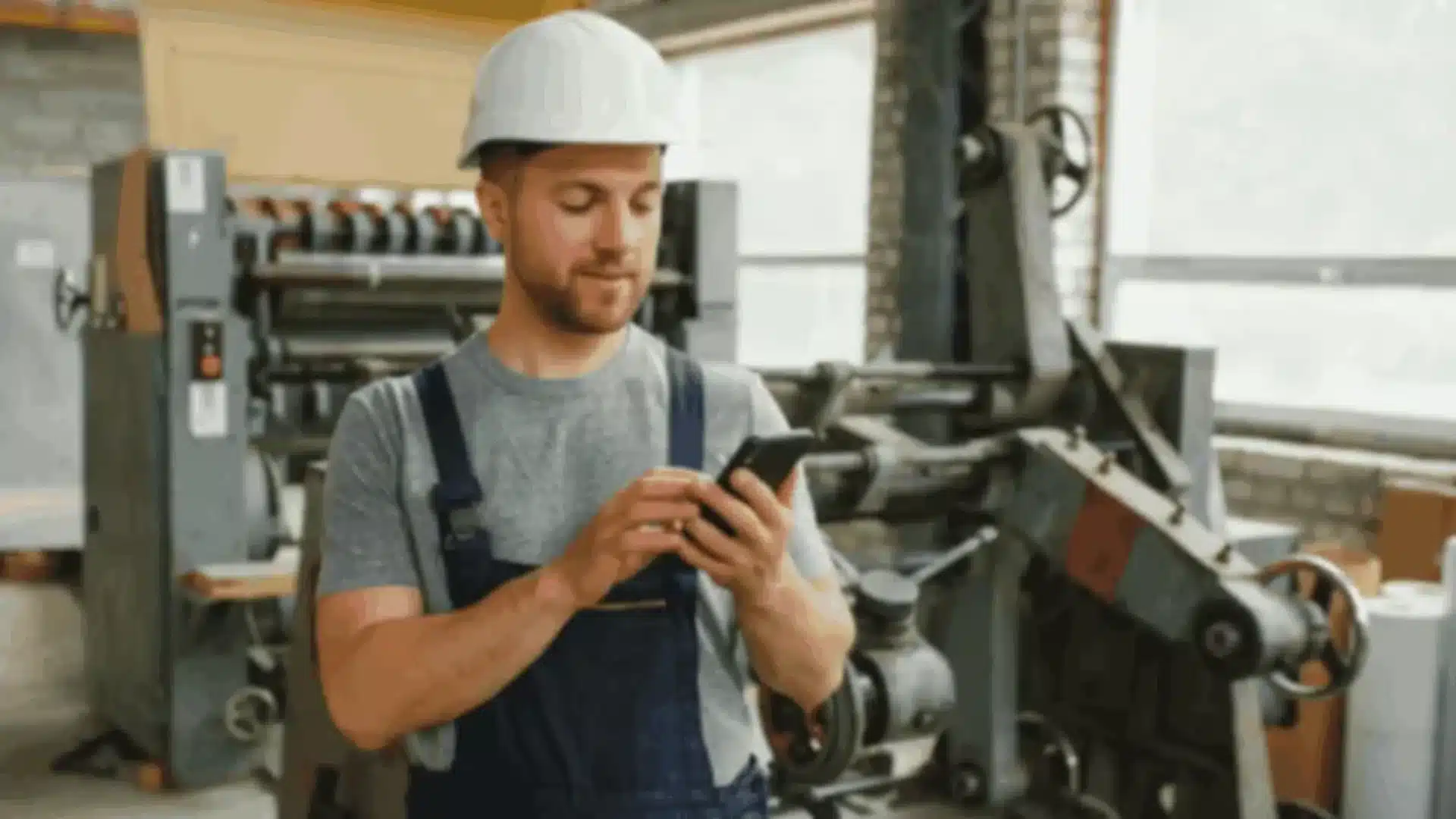 A male manufacturing worker wearing a hard hat and overalls, using a smartphone in a factory, representing the application of React Native in manufacturing app development.