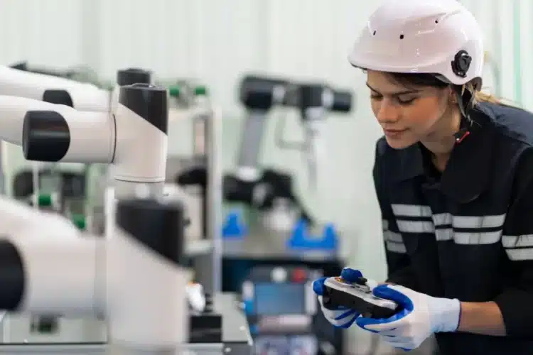 A female engineer in a safety helmet using a control device to operate robotic machinery, showcasing the role of an Android app for manufacturing in automating production processes.