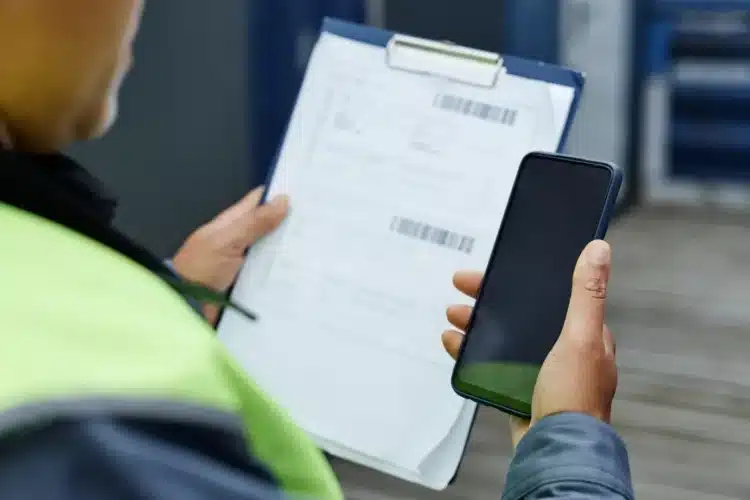 A logistics worker using an Android app for logistics on a smartphone while checking shipment details on a clipboard, streamlining delivery and tracking operations.