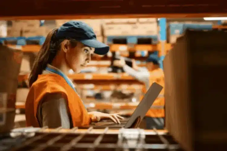 A warehouse worker wearing a vest and cap, using a laptop in a warehouse aisle with shelves of boxes, illustrating Javascript logistics app development.