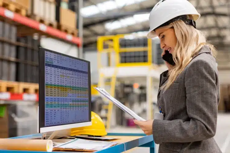 A female engineer in a hard hat reviewing data on a computer dashboard in a factory, showcasing industrial web application UI for efficient operations management.