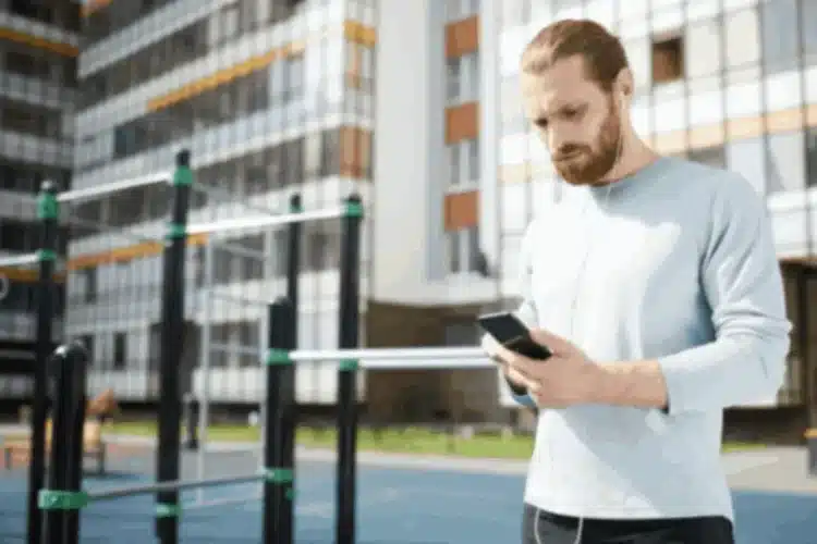 Man with earbuds checking his phone at an outdoor gym, focusing on Mobile App Quality Assurance.
