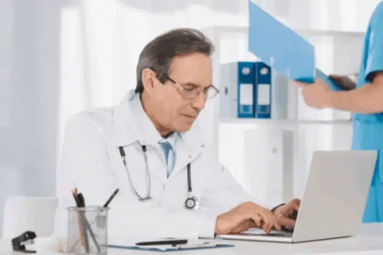 Doctor using a laptop while a nurse handles files, representing HIPAA Compliance Verification.