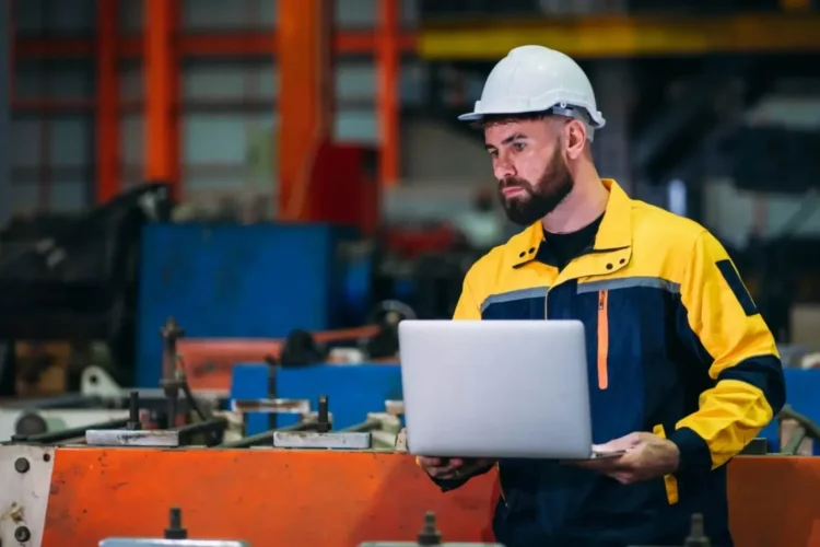 A focused engineer in a hard hat uses a laptop in a factory, showing a Manufacturing SaaS MVP.