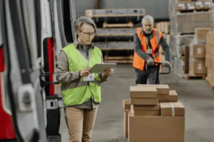 Woman in safety vest checking tablet, man moving boxes in warehouse. Supply Chain Quality Assurance.