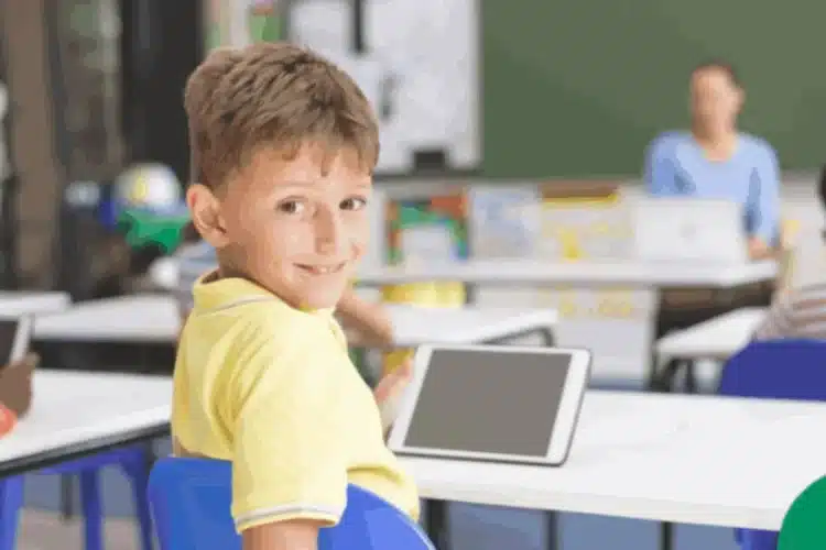 A smiling young boy in a classroom holds a tablet. Teacher and other students blurred in back. Digital Courseware Verification