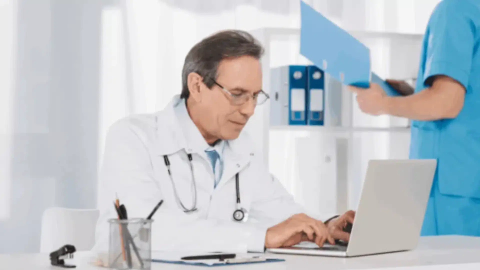 Doctor using a laptop while a nurse handles files, representing HIPAA Compliance Verification.
